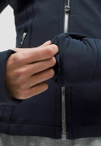 Jūras zila jaka ar gludu, elastīgu audumu. Aprīkota ar priekšējo rāvējslēdzēju un pieguļošām piedurknēm. Rožu pielāgo piedurkni, parādot elastīgu tekstūru.