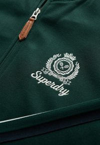 Groene sweatvest met ritssluiting, voorzien van een leren ritstrekker, geborduurd logo met kroontje en tennisontwerp, en contrasterende witte biezen.