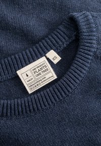 Pull en maille bleu marine avec un col côtelé. Étiquette blanche affichant la marque "NIKIN" et les instructions d'entretien, étiqueté en taille XS.