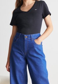 Černé tričko s v-výstřihem a krátkými rukávy, spárované s džínami modré barvy s vysokým pasem, které mají modrý pas a měděný knoflíkový detail.