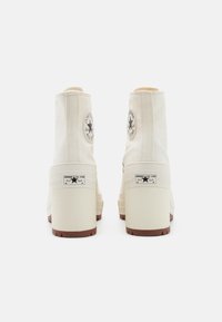 Cremfarbige Canvas-Ankle-Boots mit einem klobigen beigefarbenen Absatz, die mit dem schwarzen runden Chuck Taylor-Logo und der weißen Converse-Branding auf den Fersen versehen sind.