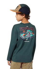 Un enfant portant un t-shirt à manches longues Timberland vert avec un graphique coloré d'un camion et de bottes dans le dos, ainsi qu'une casquette colorée.