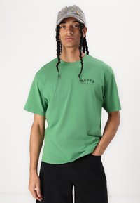 Grøn bomulds T-shirt med korte ærmer, der har en rund halsudskæring og sort grafik tekst på fronten, sammen med sorte bukser og en grå kasket.