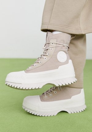 Beżowe wysokie sneakersy z prążkowaną fakturą, grubymi białymi podeszwami i sznurówkami, noszone z beżowymi spodniami na zielonym wykładzinie.