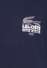 T-shirt blu navy con logo di un coccodrillo bianco e testo "LACOSTE" con l'anno "1927" sottostante, realizzato in morbido cotone.