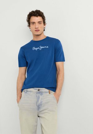 Hombre de pie frente a un fondo liso llevando una camiseta azul de Pepe Jeans, vaqueros beige claros y zapatos slip-on beige.