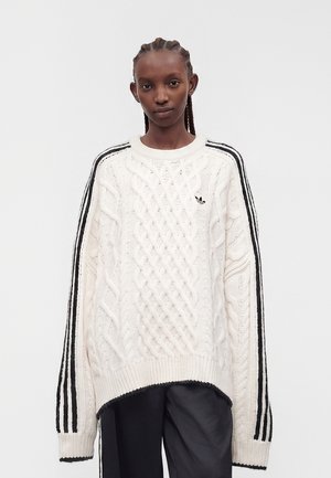 Persona con i capelli intrecciati che indossa un maglione bianco a trecce con strisce nere Adidas sulle maniche e pantaloni neri su sfondo uniforme.