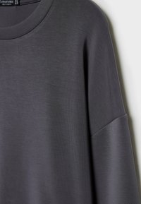 Chemise gris foncé à manches longues avec un col rond suspendue contre un arrière-plan clair.