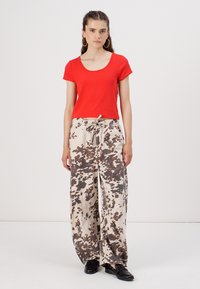 Haut ajusté rouge à manches courtes associé à un pantalon beige ample avec des motifs de éclaboussures marron foncé, taille à cordon de serrage et chaussures noires.