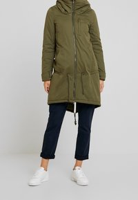 Grön parkasjacka med dragkedja fram, huva och justerbart midjeband. Har två framfickor och är kombinerad med marinblå byxor och vita sneakers.