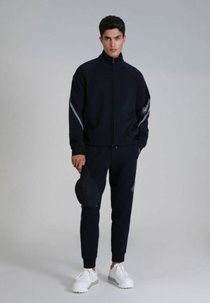 Navy tracksuit with zip-up jacket and elastic waistband pants. White stripes along sleeves, black cap held in hand, and white sneakers.