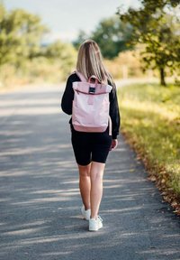 Růžový batoh s rolovacím víkem, uzaviráním na klip, nastavitelnými popruhy a hladkým povrchem, nošený osobou na stezce vedle zeleně.