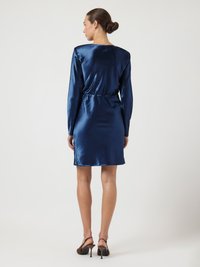 Szatén tengerészkék ruha hosszú ujjakkal, derékvonallal és egyenes szabással. Barna magas sarkú cipővel viselve, sima, fényes textúrával.