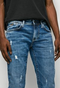 Blå denimjeans med slitna detaljer, som har fem fickor, dragkedja och en mellan-ton med subtil blekning.