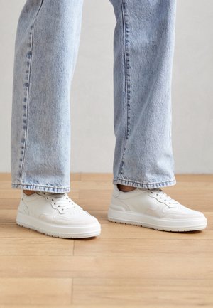 Osoba nosící světle modré džíny s rovnými nohavicemi a bílé nízké tenisky stojící na dřevěné podlaze.