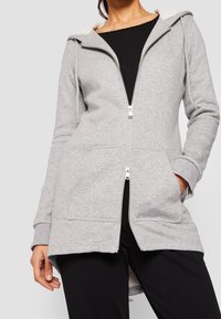Femme portant un zip-up hoodie gris clair avec des poches avant sur un t-shirt noir, les mains détendues et l'une dans la poche.