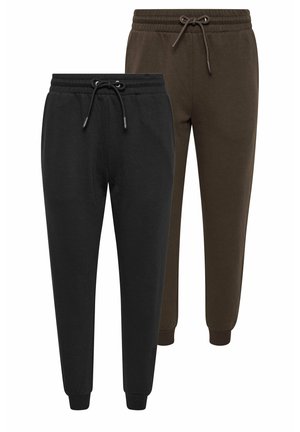 Vrouw staat, draagt zwarte joggingbroek, zwarte jas over de schouders gedrapeerd, wit T-shirt en zwarte sneakers tegen een effen lichte achtergrond.