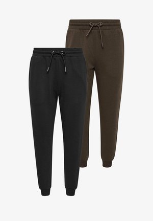 Vrouw staat, draagt zwarte joggingbroek, zwarte jas over de schouders gedrapeerd, wit T-shirt en zwarte sneakers tegen een effen lichte achtergrond.