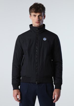Giacca nera con zip e colletto alto, dotata di una patch logo frontale, tasche laterali e polsini e orlo a coste. Tessuto dalla texture liscia.