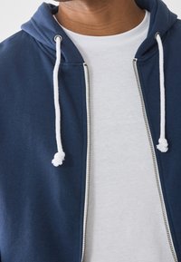 Námořnicky modrá mikina na zip s bílými šňůrkami a kovovým zipem, nošená přes bílé tričko. Měkká látka s hladkou texturou.