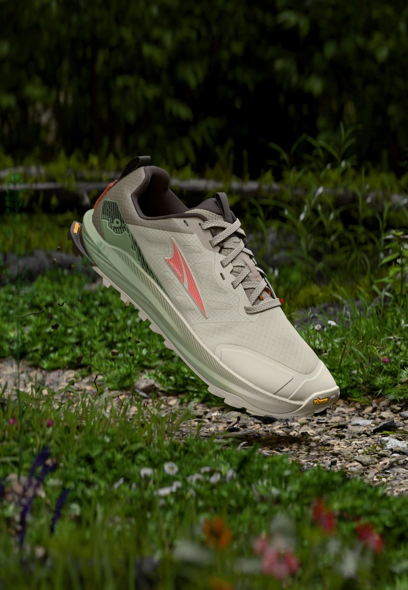 Chaussure de trail beige et verte avec un logo rose, flottant au-dessus d'un chemin de gravier entouré d'herbe verte et de fleurs sauvages.