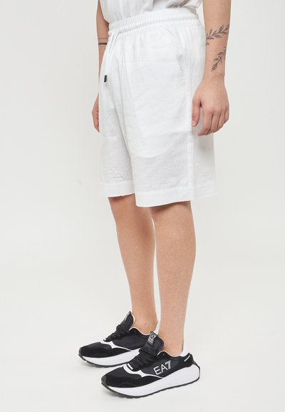 Pantaloni corti bianchi con elastico in vita, dotati di tasche laterali e una texture sottile, abbinati a sneakers bianco e nere con un logo sulla linguetta.