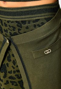 Primo piano di un maglione a maglia verde oliva con stampa leopardata nera, una cintura con strisce e un piccolo dettaglio logo metallico su una tasca.