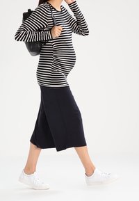 Gravid person går, klädd i en svart-vit randig långärmad tröja, svart kjol, vita sneakers och bär en svart ryggsäck.