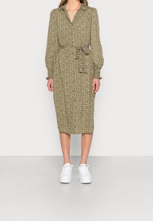 Femme portant une robe midi vert olive avec un motif floral blanc, manches longues, ceinture à la taille et baskets compensées blanches, debout sur un fond uni.