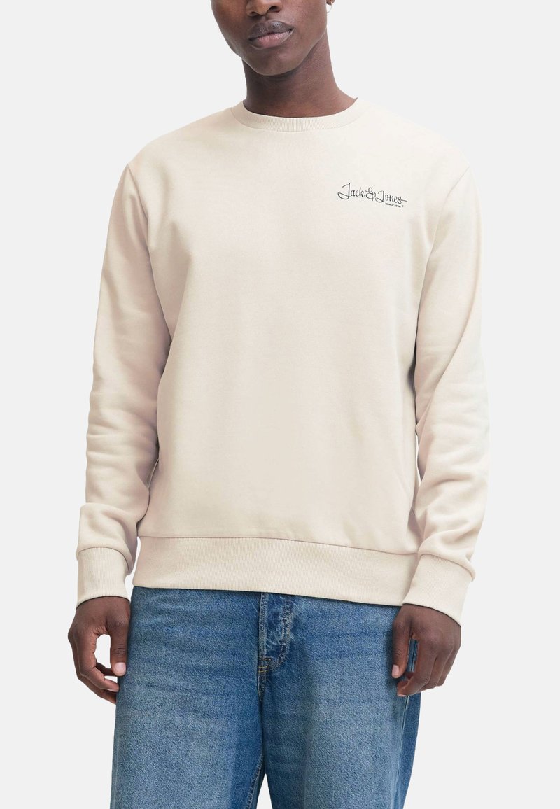 Uomo che indossa una felpa beige a maniche lunghe con il logo "Jack & Jones" sul petto e jeans di denim blu, in piedi contro uno sfondo bianco.