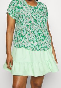 Grønne mønstrede top med korte ærmer lagdelt over en lys mintfarvet, flagrende nederdel. Har en flæset kant og åndbart stof.