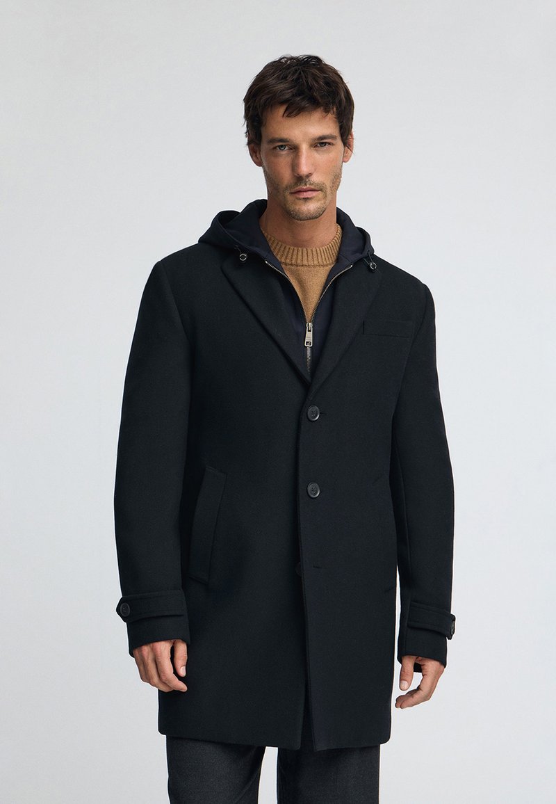 Svart överrock i ullblandning med huva, försedd med knappstängning, sidofickor och uppvikta ärmslut, lager-på-lager över en tanfärgad stickad tröja.
