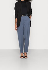 Kobieta w czarnym żakiecie, czarnym topie, niebieskich spódnicach z wysokim stanem z czarnym paskiem i czarnych sandałach na obcasie na tle białej ściany.