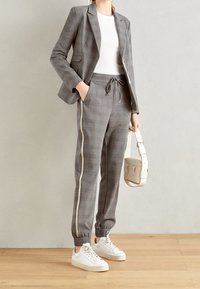 Grårutig blazer och matchande joggare med elastisk midja, med en gul sidlist. Modellen håller en krämfärgad handväska. Vita sneakers.