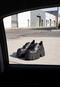 Paire de chaussures à plateforme noires épaisses avec lacets et anneaux en métal placées sur un trottoir en ville vues à travers la fenêtre d'une voiture.
