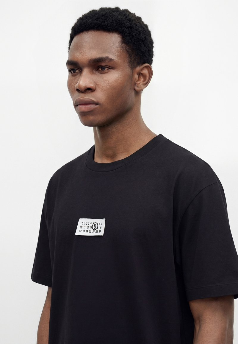 Young man with short curly hair wearing a black t-shirt with a small white printed label featuring numbers on the chest.