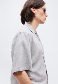 Νέος άνδρας με γκρίζο κοντομάνικο πουκάμισο και γυαλιά ηλίου, εμφανιζόμενος σε προφίλ, μπροστά σε απλό ανοιχτό φόντο.