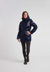 Marineblå pufferjakke med en skinnende overflade, høj krave, lynlås foran og syninger. Bæres over sorte leggings og fodtøj.