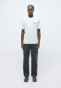 Young man with braided hair stands wearing a white Dickies T-shirt, dark gray pants, and brown shoes against a plain light background.