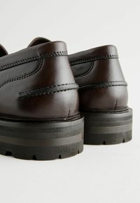Zapatos de cuero marrón oscuro con forma redondeada, parte superior texturizada y suela de goma negra gruesa con rayas horizontales.