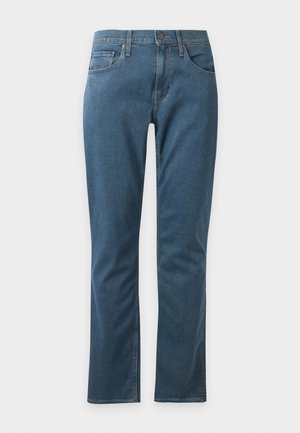 Homme portant des lunettes de soleil, une chemise beige à manches courtes boutonnée, un jean bleu et des baskets marron, debout devant un fond uni.