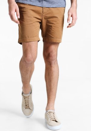 Mężczyzna w brązowych szortach podwiniętych, szarej koszulce i beżowych trampkach idący na białym tle.