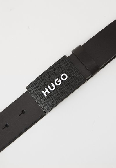 Ceinture en cuir noir avec une boucle noire texturée portant le mot "HUGO" en lettres blanches. La lanière comprend plusieurs trous de réglage.