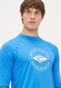 Μπλε μακρυμάνικο rash guard φτιαγμένο από ελαστικό υλικό με στρογγυλό λευκό λογότυπο που εμφανίζει "Rip Curl Surf Co" και "Established 69."