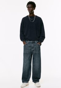 Hanorac supradimensionat bleumarin, blugi largi din denim închis, coliere argintii și adidași albi. Design minimalist cu o croială lejeră.
