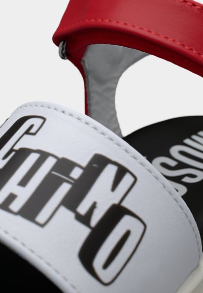 Κοντινό πλάνο λουριού σανδαλιού σε λευκό και κόκκινο με έντονο μαύρο λογότυπο «Moschino» σε λευκό φόντο.