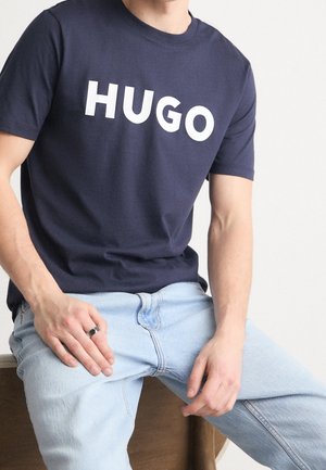 Mann trägt ein dunkelblaues T-Shirt mit "HUGO" in weißen Buchstaben und hellblauen Jeans, sitzt mit den Händen auf den Oberschenkeln.