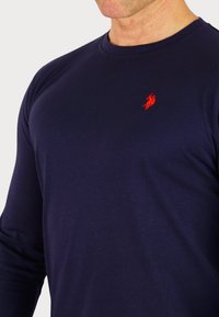 Marineblaues Langarm-T-Shirt aus Baumwolle; verfügt über ein kleines rotes gesticktes Logo auf der Brust, hat eine glatte Textur und einen klassischen Rundhalsausschnitt.