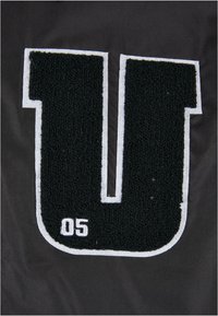 Tejido negro con una gran letra "U" texturizada en negro con contorno blanco, y "05" en blanco debajo, mostrando un diseño audaz y atlético.