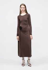 Καφέ μακρυμάνικο φόρεμα με εφαρμοστή γραμμή, διακοσμημένο με ρίγες στο πλάι και υφασμάτινο λουλούδι, συνδυασμένο με μαύρες σανδάλες με λουράκια.